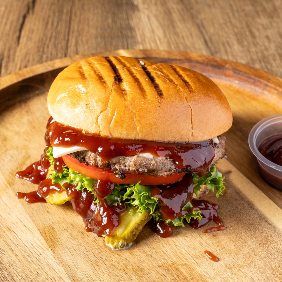 A grilled burger with melted cheese, lettuce, tomato, pickles, and barbecue sauce on a wooden board.