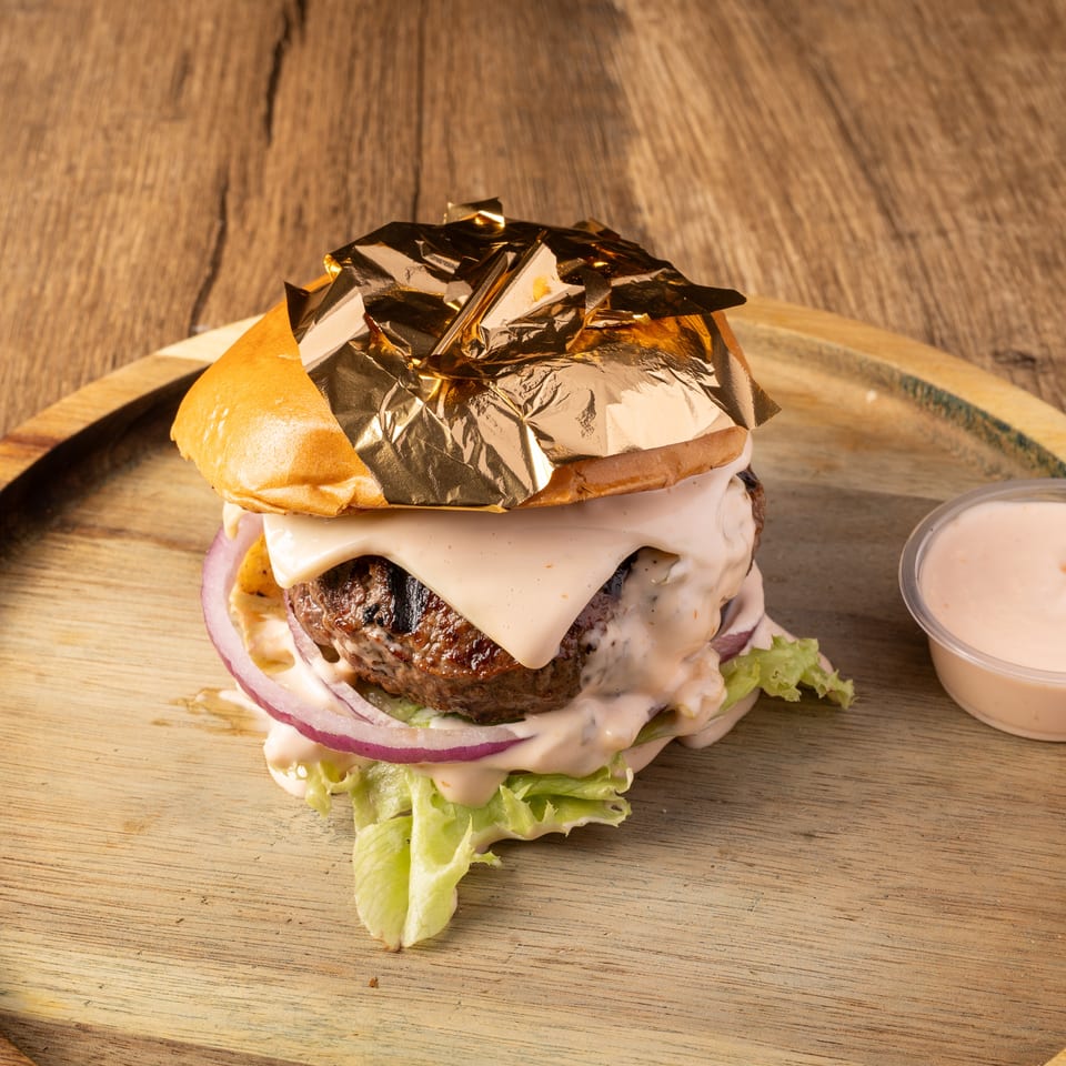 Gourmet burger with gold leaf, cheese, lettuce, red onion, and sauce on a wooden tray with a side of sauce.