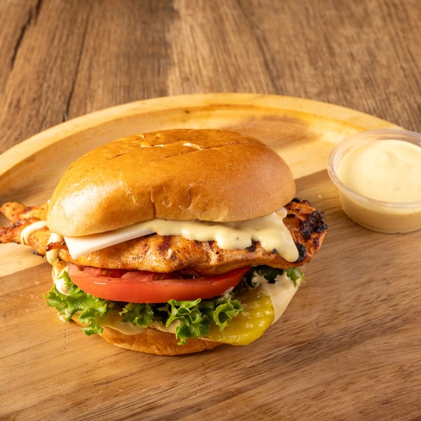 Grilled chicken sandwich with cheese, lettuce, tomato, and pickles, served with a side of sauce.