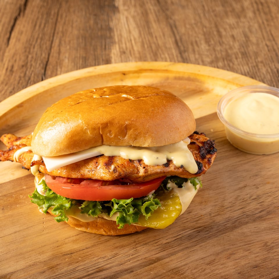 Grilled chicken sandwich with cheese, lettuce, tomato, and pickles, served with a side of sauce.