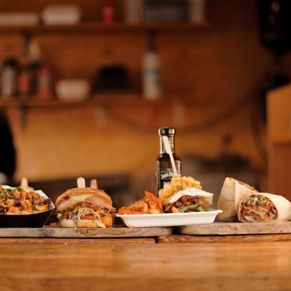 A wooden counter displays various dishes including loaded fries, burgers, and wraps, with a chef in the background.