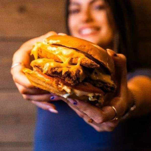 Hands holding a large, cheesy double burger with tomato, extended forward.