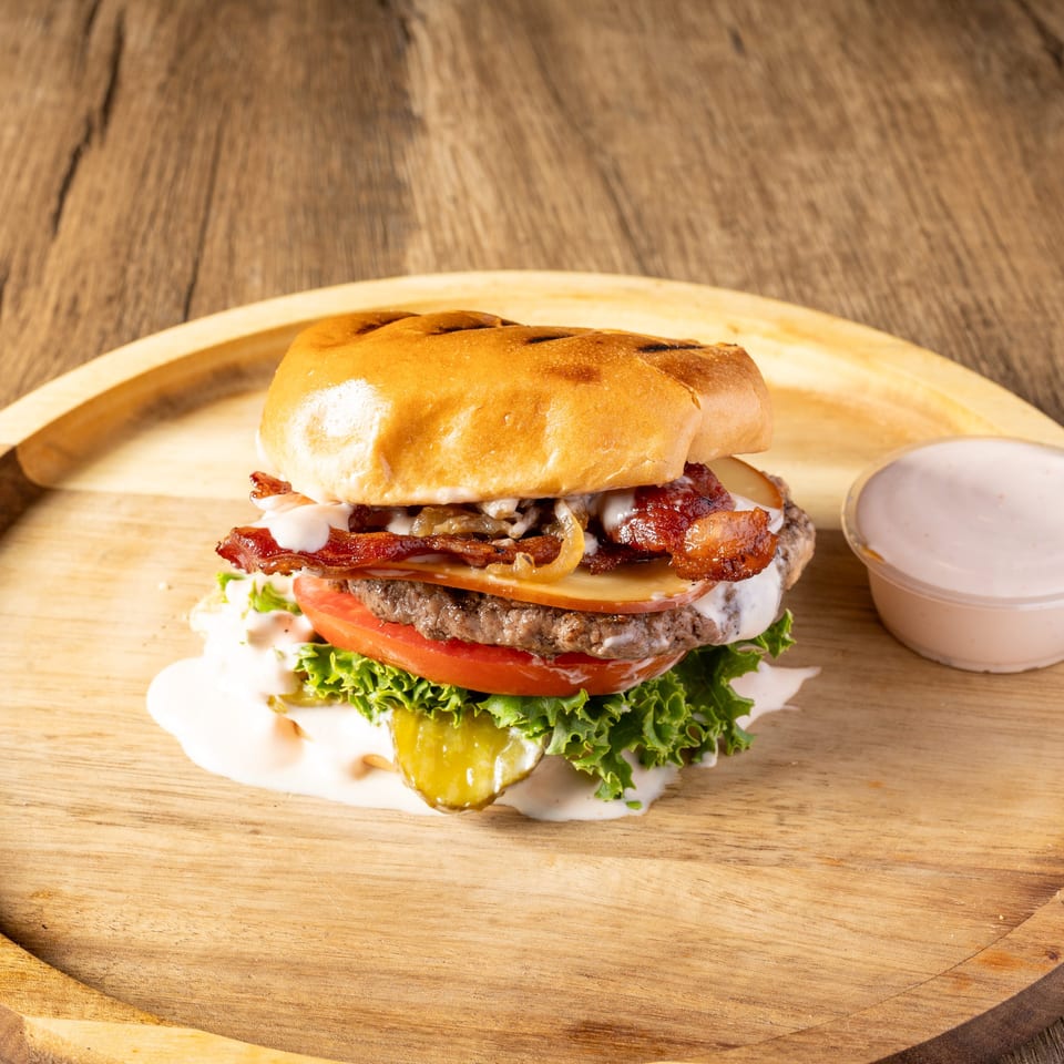 A gourmet burger with bacon, cheese, lettuce, tomato, and sauce on a wooden plate.