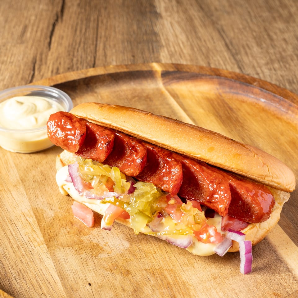 Grilled hot dog in a bun with relish, onions, and sauce, served on a wooden plate.