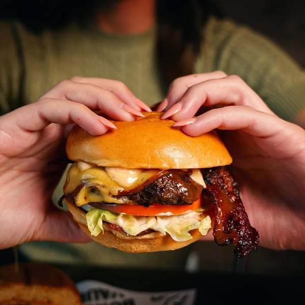 Hands holding a gourmet burger with melted cheese, crispy bacon, lettuce, and tomato.